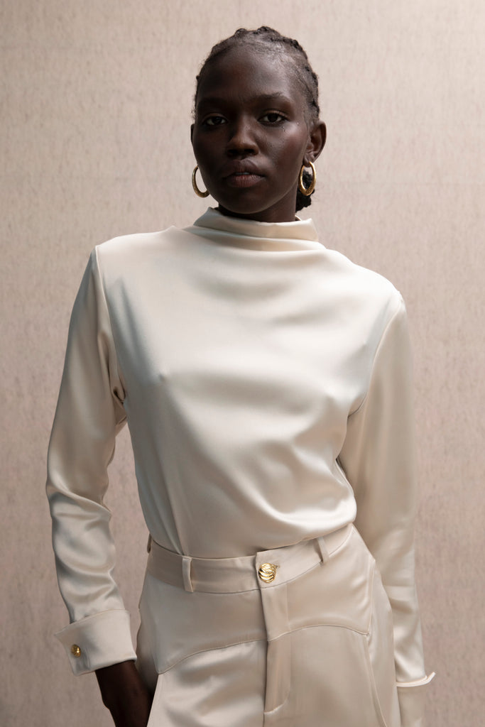 Champagne Vivien LS Top Elevate your wardrobe with this silky-soft and versatile satin top.-Cowl neckline with stacked collar-Self-loops with gold knot buttons at back neck-Removable shoulder pads with interior pockets-Hidden zip at back-Turnback cuff with gold knot button-Fluid drape with tailored structure-Finished with French seamsWe recommend ordering your usual size.Styled with the Vivien Pant & Ava Skirt