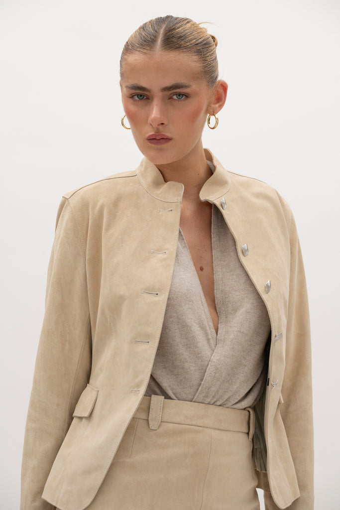 Sand Hyde Jacket -Structured vintage inspired jacket with mandarin collar-Princess seams contour the bodice and waist -100% calf leather (suede) -Ornate front button hardware with interior stay buttons for durability-Hidden zipper pockets integrated into front princess seams-Buttoned pocket flaps for visual detail (non-functional flap; real pocket hidden beneath)-Clean-finished hem, sleeve, and collar with subtle edge stitch detailing-Hidden interior chest pocket with zipper closure-Vents at back of sleeves-Fully linedWe recommend ordering your usual size.Styled with the Meredith Skirt